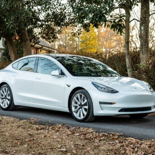 Tesla Model 3 parked on a forested driveway photographed from the passenger side front quarter panel.