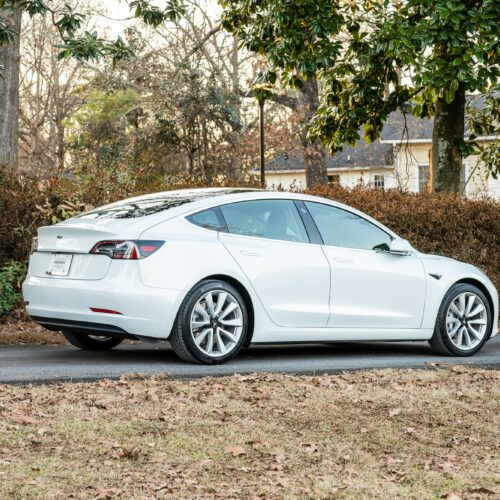 Tesla Model 3 parked on a forested driveway photographed from the passenger side rear quarter panel.