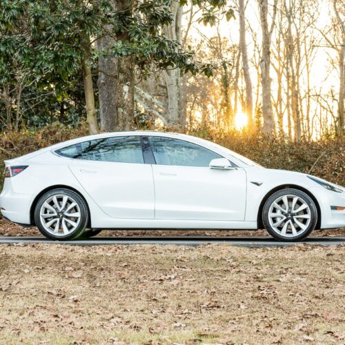 Tesla Model 3 parked on a forested driveway photographed from the passenger side.