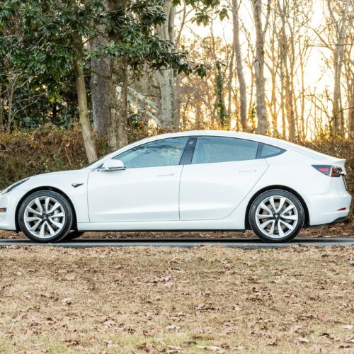 Tesla Model 3 parked on a forested driveway photographed from the driver side front quarter panel.