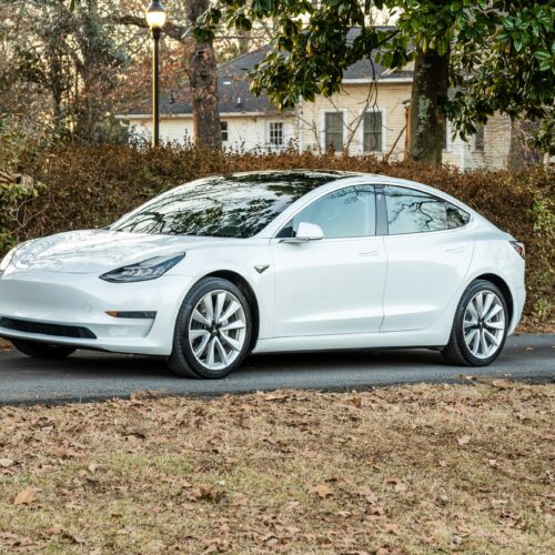 Tesla Model 3 parked on a forested driveway photographed from the driver side front quarter panel.