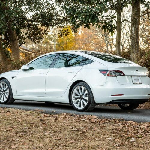 Tesla Model 3 parked on a forested driveway photographed from the driver side rear quarter panel.