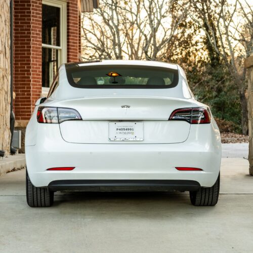 Tesla Model 3 parked on a forested driveway photographed from the rear.